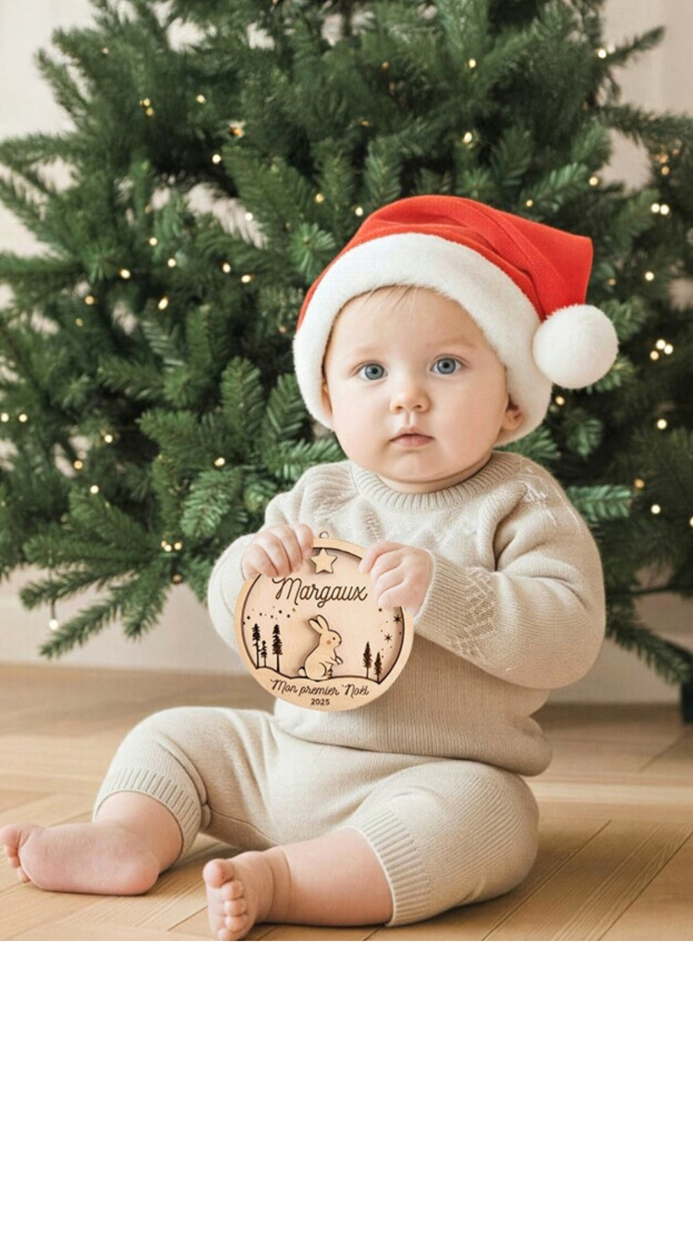 Bébé assis devant un sapin tenant une boule de Noël personnalisée en bois, ambiance douce et naturelle