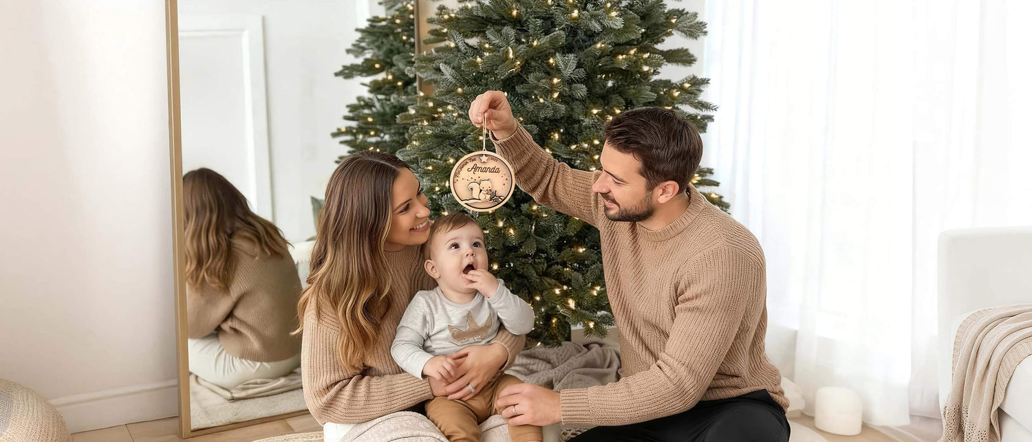 Famille devant le sapin tenant une boule de Noël personnalisée en bois pour le premier Noël de bébé