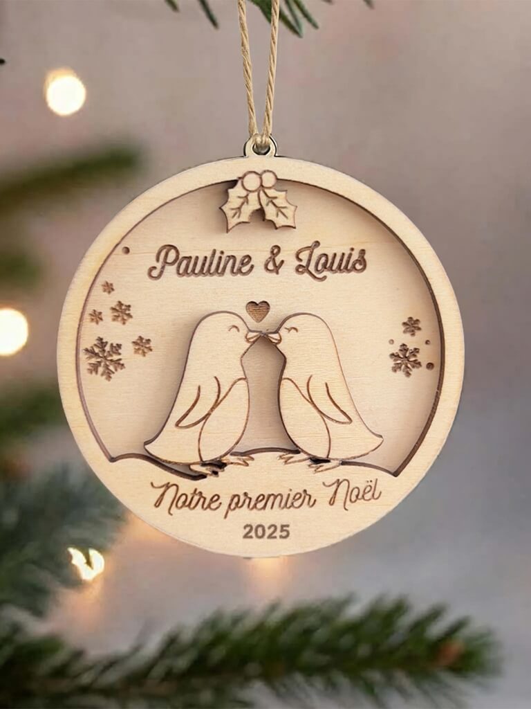 Boule de Noël en bois personnalisée motif Couple de Pingouins, suspendue dans un sapin décoré.